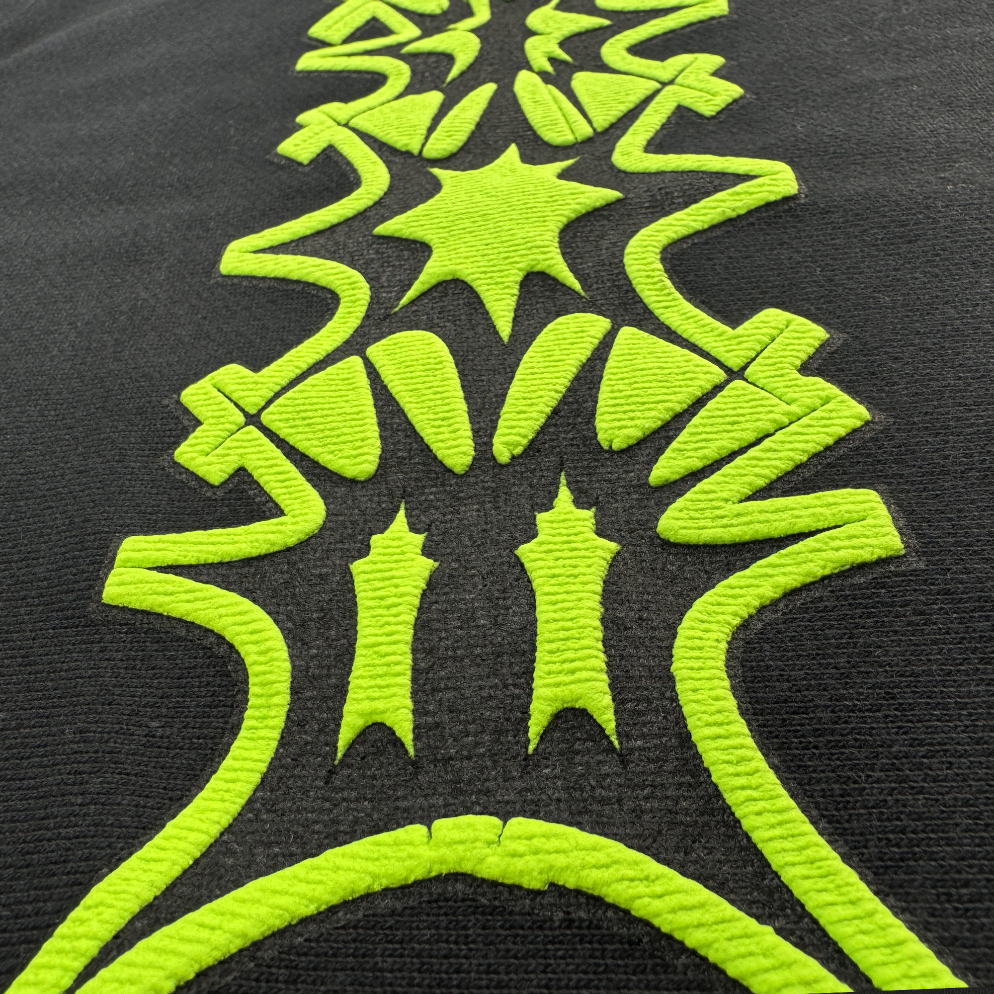 The BOSK Distortion Hoodie features an intricate black fabric design with vivid green embroidery. It showcases a symmetrical geometric pattern of abstract shapes, star-like forms, and curved lines arranged vertically. An oversized drop shoulder silhouette adds contrast to the dark background.
