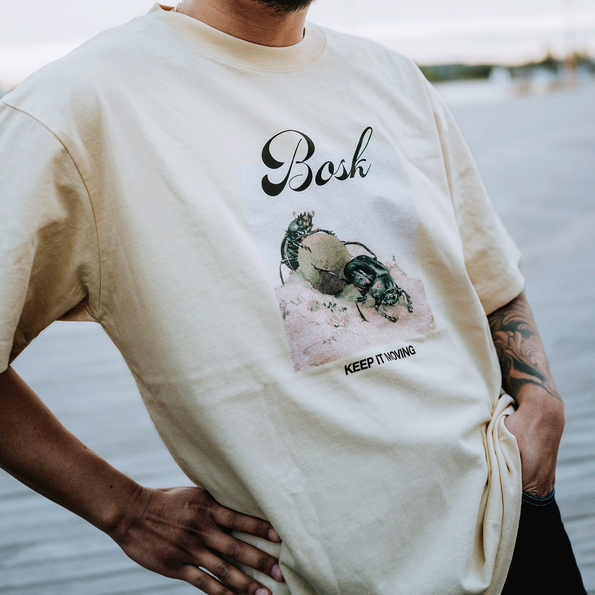 Wearing the BOSK Moving Tee, a classic-fit heavy-weight cotton shirt with two beetles pushing a ball, “BOSK” on top and "KEEP IT MOVING" below, a person stands outdoors on a boardwalk with one hand on their hip.