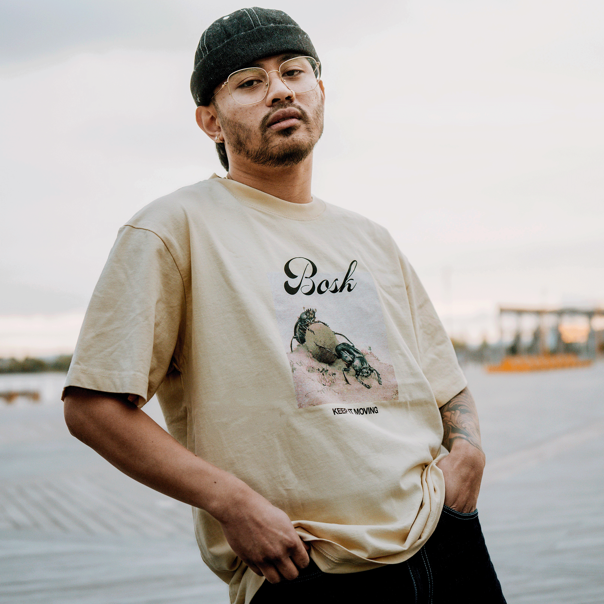 A man in glasses and a black cap stands on a boardwalk, holding his beige BOSK Moving Tee—100% cotton with a crab graphic and "KEEP MOVING" text—against a blurred outdoor backdrop of sky and distant structures.