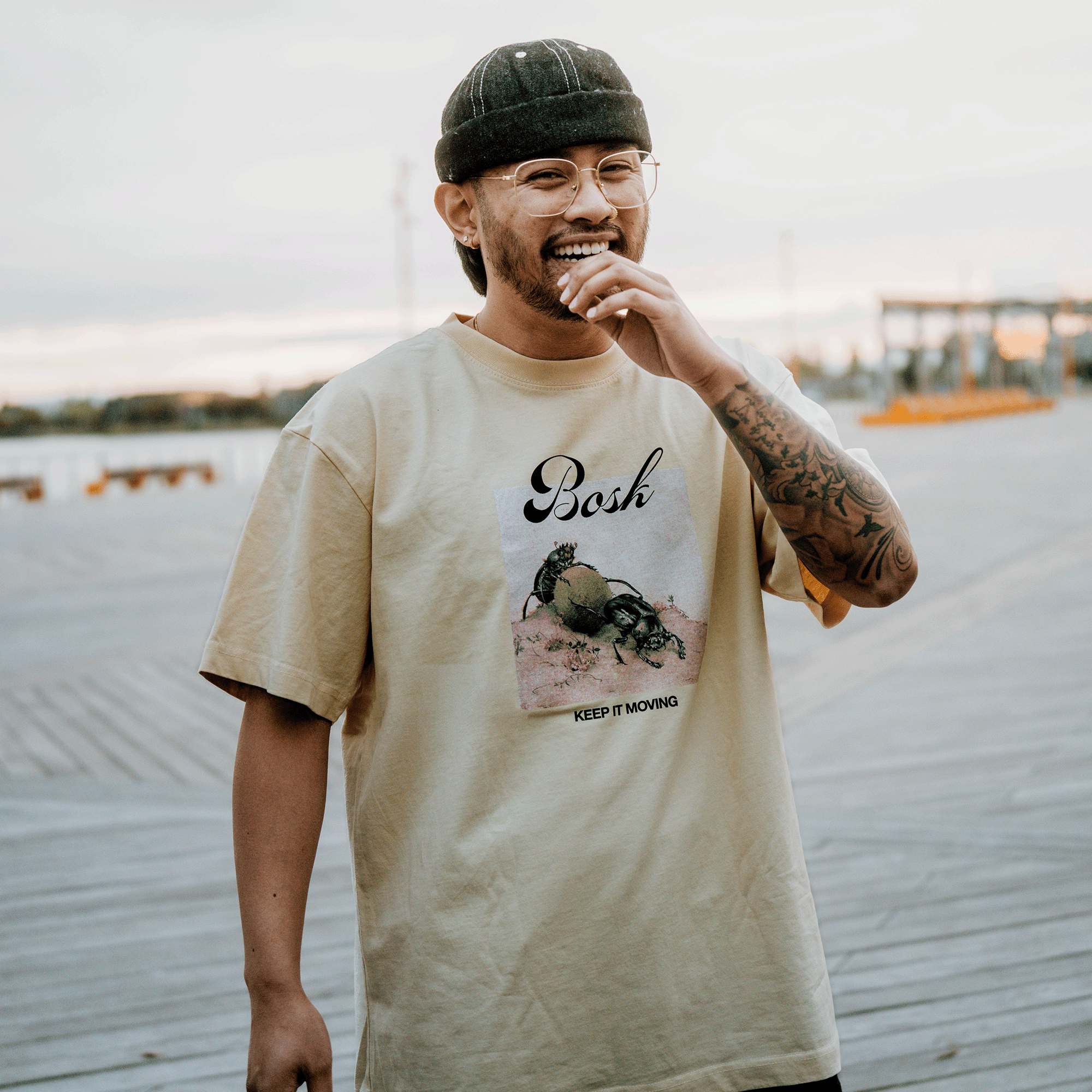 A smiling, bearded man with glasses stands on a boardwalk in the BOSK Moving Tee—a classic fit, off-white, heavy-weight cotton shirt featuring a crab graphic and “Bosh KEEP IT MOVING” text. Tattoos show against the cloudy sky and outdoor structures.